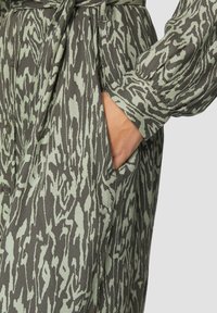 Mustriga kleit, mis on valmistatud hallrohelisest kangast lainelise mustriga, nuppudega detailide ja küljetaskuga. Vöökoht on rõhutatud ning varrukate otstes on pompoonid.