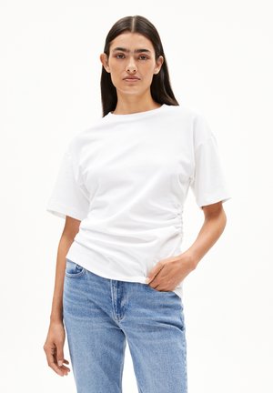 White cotton t-shirt with a relaxed fit, short sleeves, and gathered side detail, paired with light blue denim jeans.