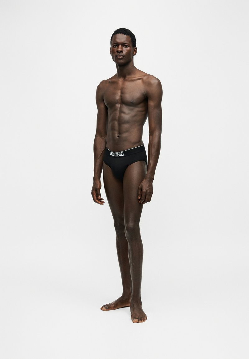 Slanke donkerhuidige man staat blootsvoets tegen een effen achtergrond, gekleed in zwarte Diesel boxers met logo op de tailleband.