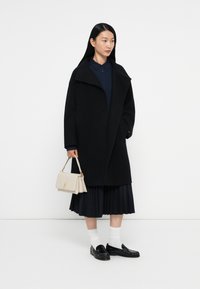 Zwarte wollen jas met oversize kraag, gedrapeerde silhouet, gedragen over een marineblauwe shirt. Gepinde zwarte rok en zwarte loafers; beige handtasdetails zichtbaar.