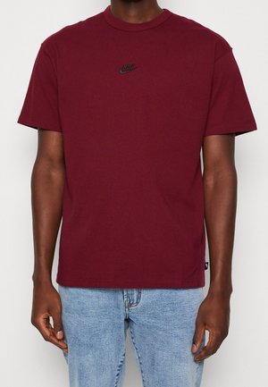 Mand iført en bordeaux Nike T-shirt med et lille sort logo på brystet og lyseblå jeans, der står foran en ensfarvet baggrund.