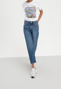 Vit t-shirt med Claude Monet-motiv, blå högmidjade jeans och vita sneakers. Löst sittande, rak ben-design, lätt denim-textur.