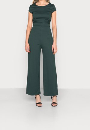 Femme portant une combinaison short manches courtes vert foncé avec jambes larges et des sandales à talons ouvertes noires, debout contre un mur clair.