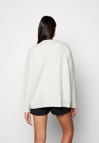 Světle šedý oversized svetr s kulatým výstřihem, spadenými rameny a vroubkovanými manžetami. Pletená textura, nošený v převisu přes tmavé šortky.