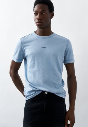 Νέος άνδρας που φοράει ανοιχτό μπλε μπλουζάκι BOSS και σκούρο παντελόνι, στέκεται με τα χέρια πίσω από την πλάτη, κοιτώντας προς το πλάι σε λευκό φόντο.