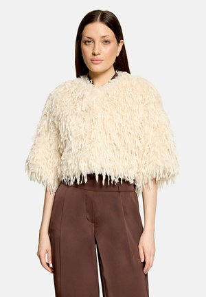 Vrouw met steil donker haar, gekleed in een crèmekleurig gestructureerd bontjasje en hooggetailleerde bruine wijde broek tegen een effen achtergrond.
