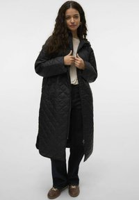 Abrigo largo negro acolchado con cremallera al frente, que cuenta con capucha y mangas anchas. Lleva puesto sobre una camisa clara y jeans oscuros, combinado con zapatos marrones.