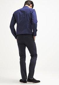 Marineblaues Shirt mit weißen Polka-Dots, aus Baumwollmischung, taillierte Passform. Dunkle Hose, gerade geschnitten, dezente Gesäßtaschen, getragen mit schwarzen Schuhen.