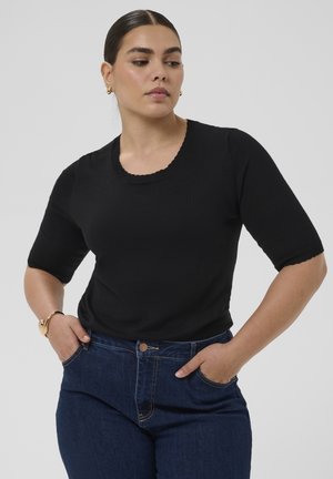 Donna con capelli scuri, indossa una maglietta nera a maniche corte e jeans blu, con le mani nelle tasche, guarda verso il basso e di lato su uno sfondo semplice.