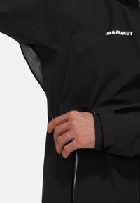 Veste noire imperméable avec un extérieur texturé, des poignets ajustables et une poche zippée sur le côté. Logo de la marque visible sur la poitrine.
