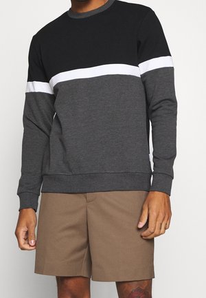 Mężczyzna ubrany w czarną, białą i ciemnoszarą bluzę z długim rękawem w blokowe kolory oraz brązowe krótkie spodenki sięgające kolan.