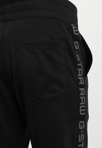 Černé tepláky s texturovaným páskem v pase, se zadní kapsou a bočním pruhem s šedým logem "G-STAR RAW".