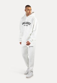Bílá mikina s kapucí s černým potiskem "SMILODOX ATHL WEAR", v kombinaci s bílými tepláky se značkou na noze. Nošeno s bílými teniskami.