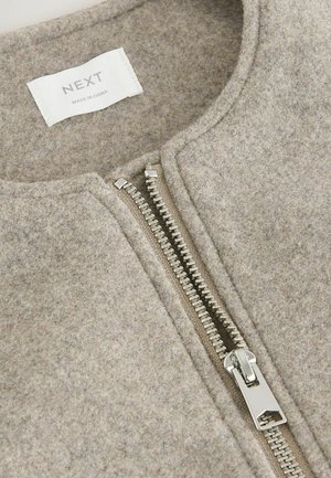 Beżowa kurtka z mieszanki wełny o gładkiej fakturze, z metalowym suwakiem w kolorze srebrnym i okrągłym dekoltem, oznaczona etykietą "NEXT" w pobliżu kołnierza.