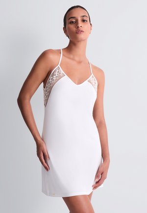 Femme portant une robe blanche sans manches avec des panneaux en dentelle près des épaules, debout devant un fond uni gris clair.