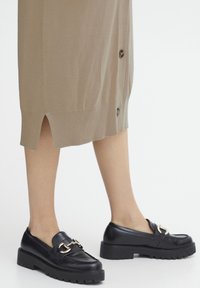 Mocassini di pelle nera con suole spesse, con un accentuato fulcro dorato. L'outfit include un vestito beige chiaro, lungo fino al ginocchio, con bottoni.