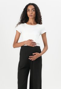 Hvit kortermet skjorte kombinert med løse sorte høytlignende bukser. Modellen poserer med hendene hvilende på midje, og viser frem et enkelt design.