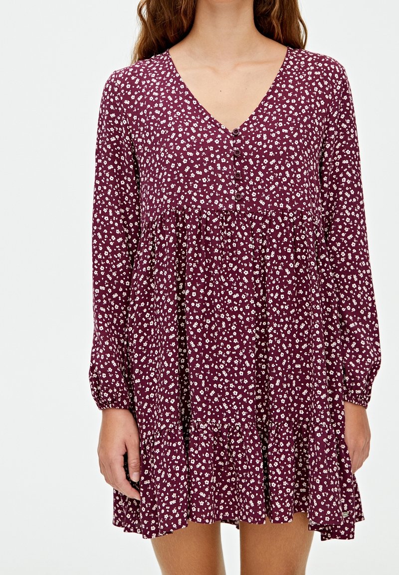 Robe bordeaux avec col en V, manches longues et motif floral blanc. Comprend une jupe à volants avec un ourlet à volants et des détails à boutons sur le devant.