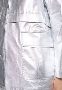 Gros plan sur une poche de veste argentée métallique avec détail de rabat, coutures visibles et une partie du bras d'une personne portant la veste.
