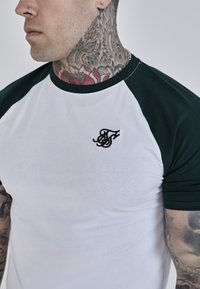 Bijela majica s zelenim raglan rukavima i malim crnim izvezenim logom na prsima. Ima prilagođeni siluetu i glatku teksturu.