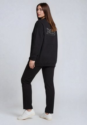 Nuori nainen seisoo, päällään musta collegepaita, jossa strassikirjaimet selässä, mustat housut ja valkoiset tennarit harmaalla taustalla.