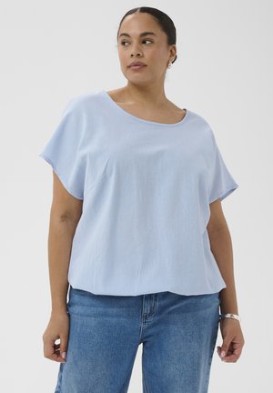 Žena má na sebe svetlomodré tričko s krátkym rukávom a modré džínsy, stojí pred jednoduchým bielym pozadím a pozerá sa doľava.