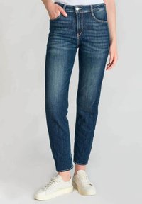 Jeans skinny de color azul oscuro con una textura suave, que cuentan con un cierre de botón en la parte frontal, cinco bolsillos y un ligero desgaste en los muslos. Combinados con zapatillas blancas.