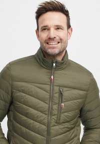Olivgrüne gepolsterte Jacke mit hohem Kragen, Reißverschluss vorne und diagonalem Steppmuster. Mit roten und weißen Reißverschlussanhängern und Seitentaschen.