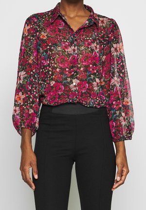 Kvinde iført en gennemsigtig langærmet knapbluse med et levende rødt, pink og orange blomsterprint, kombineret med højtaljede sorte bukser.