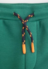 Zielone bawełniane spodnie dresowe mają elastyczny pas z czarno-pomarańczowym plecionym sznurkiem do ściągania. Materiał jest miękki, z widocznymi detalami szwów.