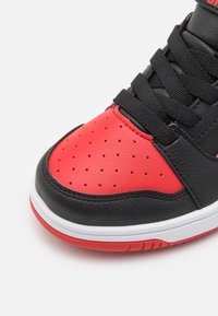 Zwarte en rode sneaker met een geperforeerde rode leren neus. Voorzien van zwarte veters en een rubberen witte tussenzool met een rode accent op de buitenzool.