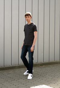 T-shirt gris foncé à manches courtes avec un petit logo "Lee", associé à un jean bleu foncé et des baskets blanches. Le modèle porte un bonnet beige.