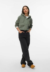Grøn corduroy hoodie med lynlås foran, to frontlommer og ribbede manchetter, parret med sorte jeans og brune slip-on sko på en hvid baggrund.