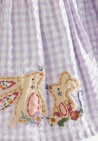 Dos animales bordados en beige, un conejo y un ratón, con detalles florales sobre una tela de cuadros púrpuras y blancos.