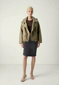 Femme portant une veste oversized marron clair, un haut cropped marron foncé, une jupe noire mi-longue et des sandales à talons transparents, debout.