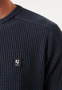 Navyfarbener Strickpullover mit strukturiertem Muster und geripptem Ausschnitt. Mit einem kleinen schwarzen Logo-Patch auf der linken Brust.