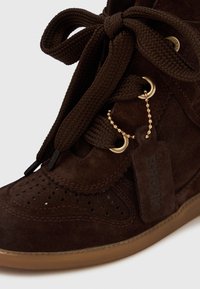 Sapatilha alta em camurça castanha com atacadores trançados, ilhós dourados, design perfurado e sola de borracha castanha clara. Apresenta uma etiqueta com a marca "BRONX".