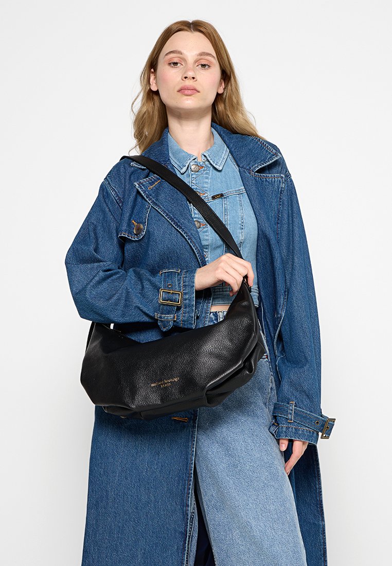 Jeune femme portant une veste en denim superposée et un jean tient un sac à bandoulière en cuir noir avec des lettres dorées devant un fond blanc.