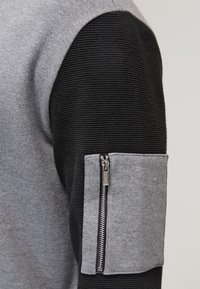 Pull gris et noir avec une texture côtelée, doté d'une poche zippée sur la manche. Le tissu est doux, alliant des motifs lisses et texturés.