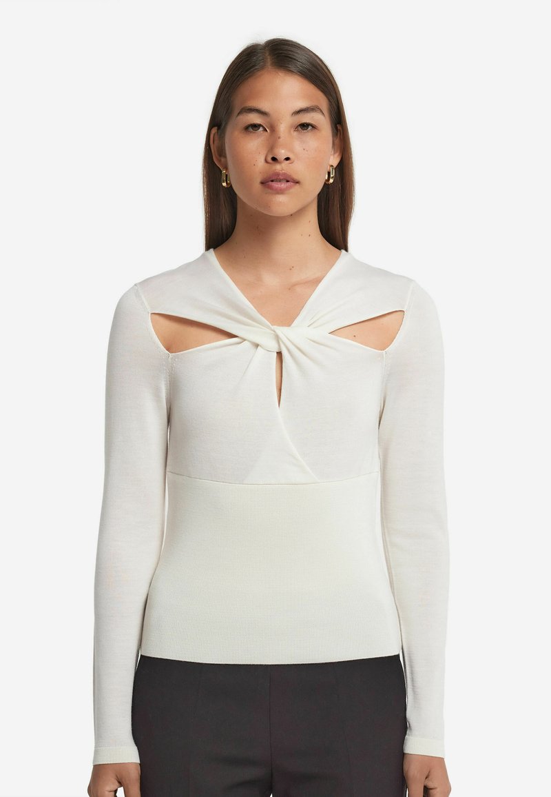 Vit långärmad topp med en vriden halsringning, utskärningar vid axlarna och en åtsittande ribbad struktur runt midjan.