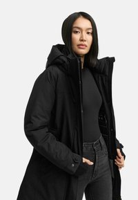 Parka nera imbottita con un grande cappuccio, chiusura con zip e tasche frontali. Il tessuto appare liscio con una finitura opaca, adatto per il freddo.