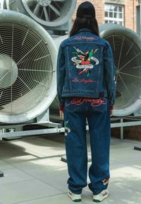 Persona vistiendo una chaqueta y pantalones de mezclilla azul a juego, con coloridos diseños de pájaros y corazones estilo tatuaje, de pie frente a grandes ventiladores industriales.