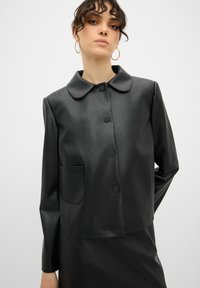 Schwarze Kunstlederjacke mit rundem Kragen, vorderem Knopfverschluss und einer einzigen Tasche auf der linken Seite. Glatte Textur und cropped Design.