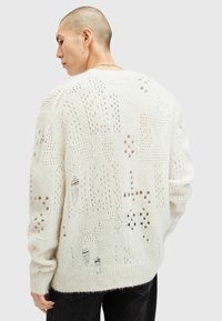 Cremefarbener Strickpullover mit Lochmustern, lockerer Passform und gerippten Bündchen. Der Rücken zeigt verschiedene Texturen und Designs.
