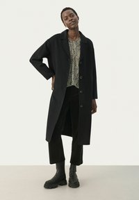 Schwarzer langer Mantel mit Reverskragen, großen Knöpfen und seitlichen Schlitzen. Kombiniert mit einer gemusterten Bluse, dunklen Cordhosen und schwarzen Stiefeln.