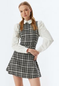 Vestido a cuadros en blanco y negro con un corsé ajustado y falda acampanada, llevado sobre una camisa blanca de manga larga con cuello.