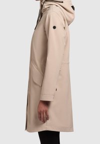 Manteau imperméable beige avec capuche, poches latérales et cordon de serrage ajustable. Il présente une texture lisse et des boutons accentués noirs.