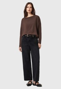 Camicia marrone a maniche lunghe con scollatura ampia, abbinata a jeans in denim neri a vita alta. Il modello indossa delle scarpe piatte nere.