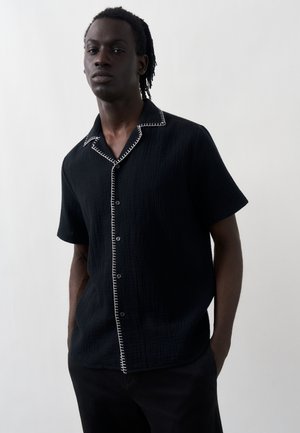 Mężczyzna z krótkimi dredami, ubrany w czarną koszulę z krótkim rękawem na guziki z białym przeszyciem wzdłuż kołnierza i przodu.