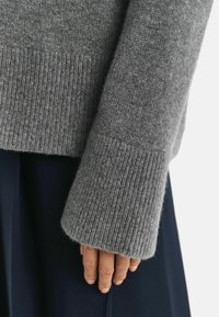 Grau gestrickter Pulloverärmel mit geripptem Bund, der eine weiche Textur zeigt. Akzente sind eine sichtbare Naht in der Nähe der Hand, kombiniert mit einem marineblauen Rock.